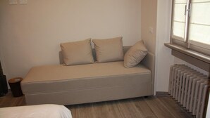 Une petite chambre avec un lit simple, un canapé-lit, un radiateur et une fenêtre avec des stores.