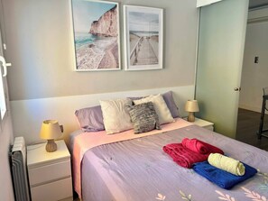 A bedroom with a bed, pillows, a nightstand with a lamp, and two framed pictures on the wall. - Oasis Sitges Apartment: Relaxing, Centrally Located, and With a Community Pool (Sitges)