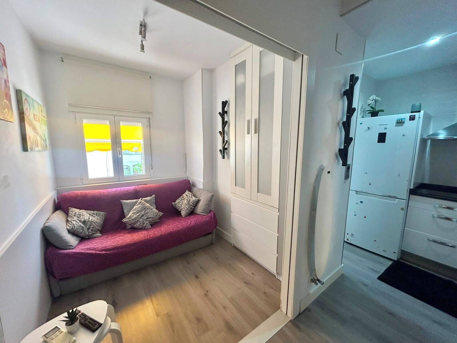 Un espacio habitable compacto con sofá, área de cocina con refrigerador y ventana con persianas.