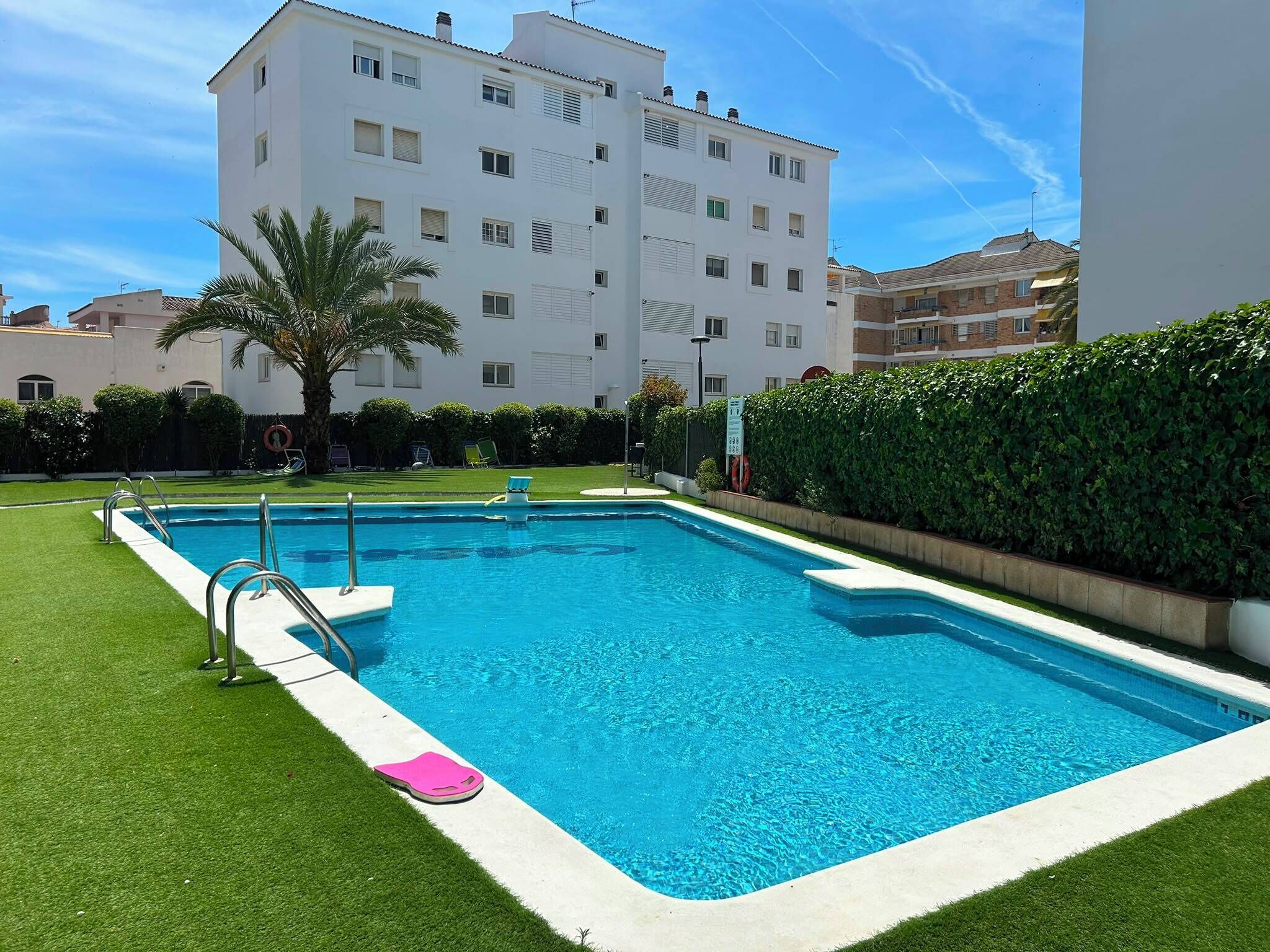 Una piscina rectangular con una escalera de metal, rodeada de césped artificial, una palmera y un seto.