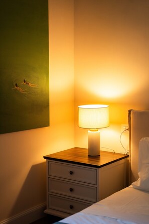 Ein ordentlich bezogenes Bett mit weißer Decke und Kissen, ein Nachttisch mit Lampe und ein grünes Bild an der Wand.