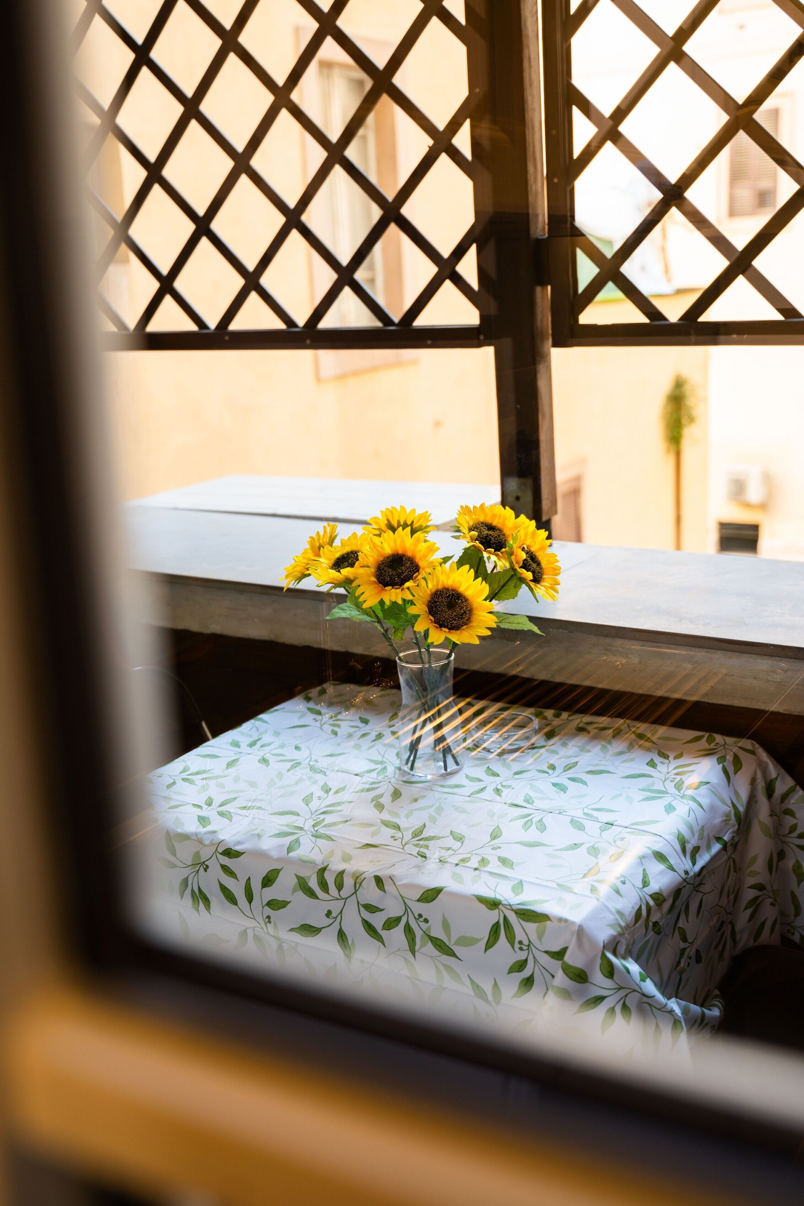 A window with a lattice design, a table with a leaf-patterned tablecloth, and a vase with sunflowers.