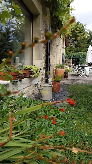 A stone building with potted plants, a metal watering can, and outdoor furniture.