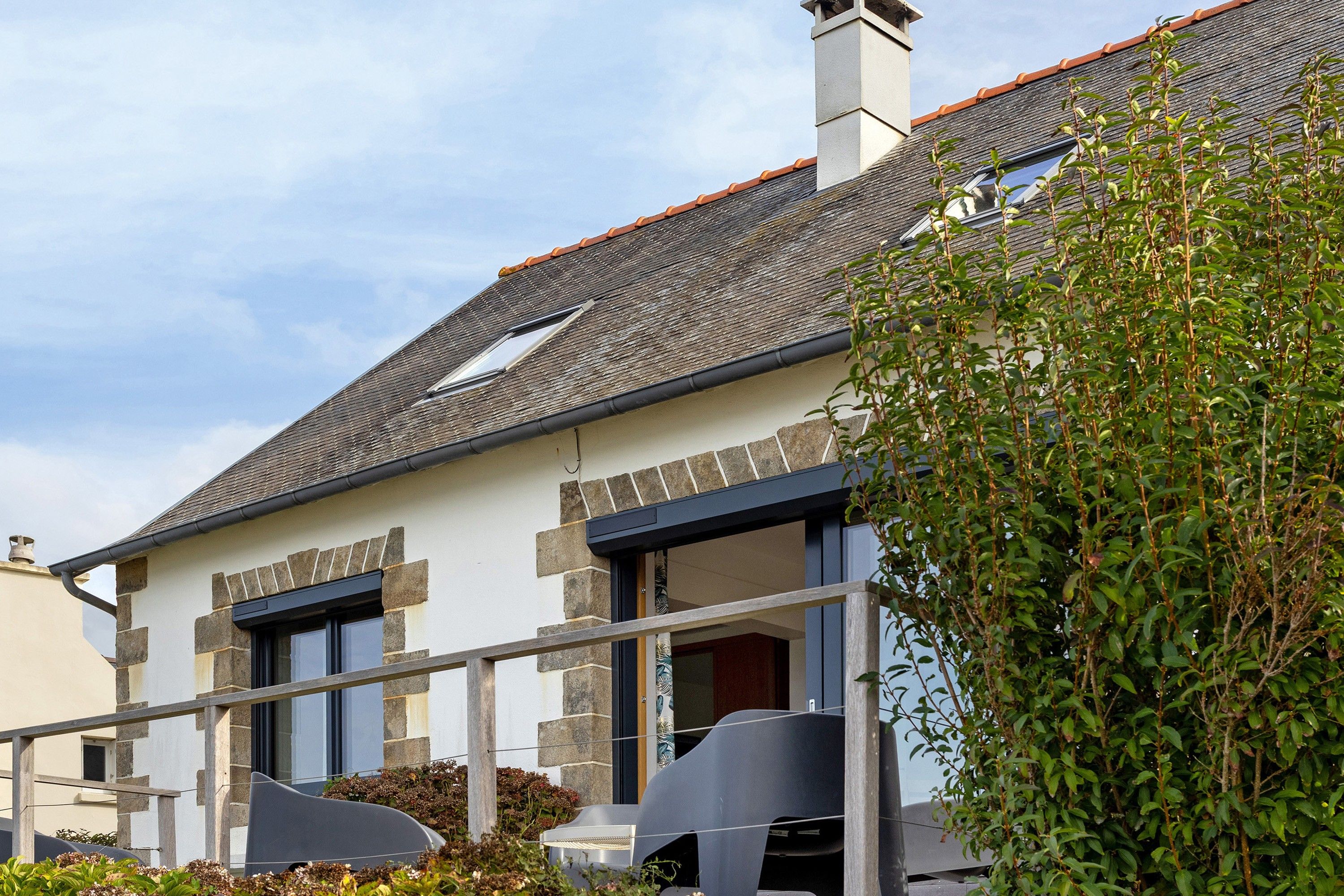 Une maison à deux étages, avec un toit en pente, une façade en pierre et une cheminée bien visible.