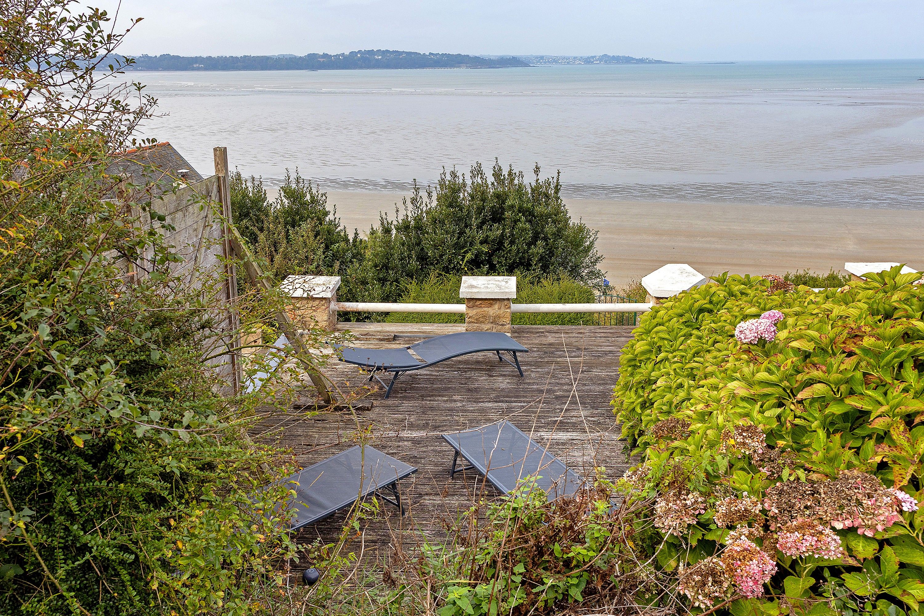 A wooden deck with two lounge chairs and a stone bench overlooking a beach.