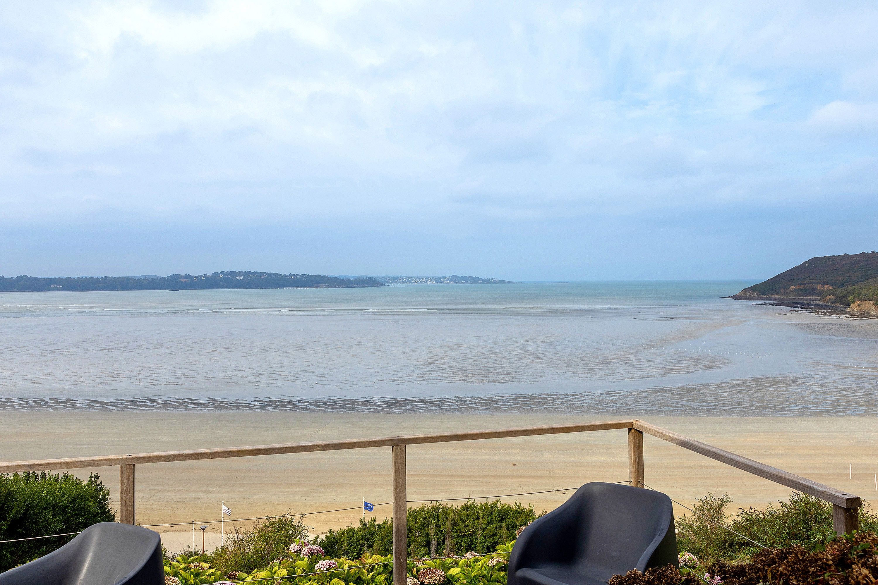 Une vue sur la plage, avec une terrasse en bois et deux chaises, donnant sur la mer et la côte lointaine.