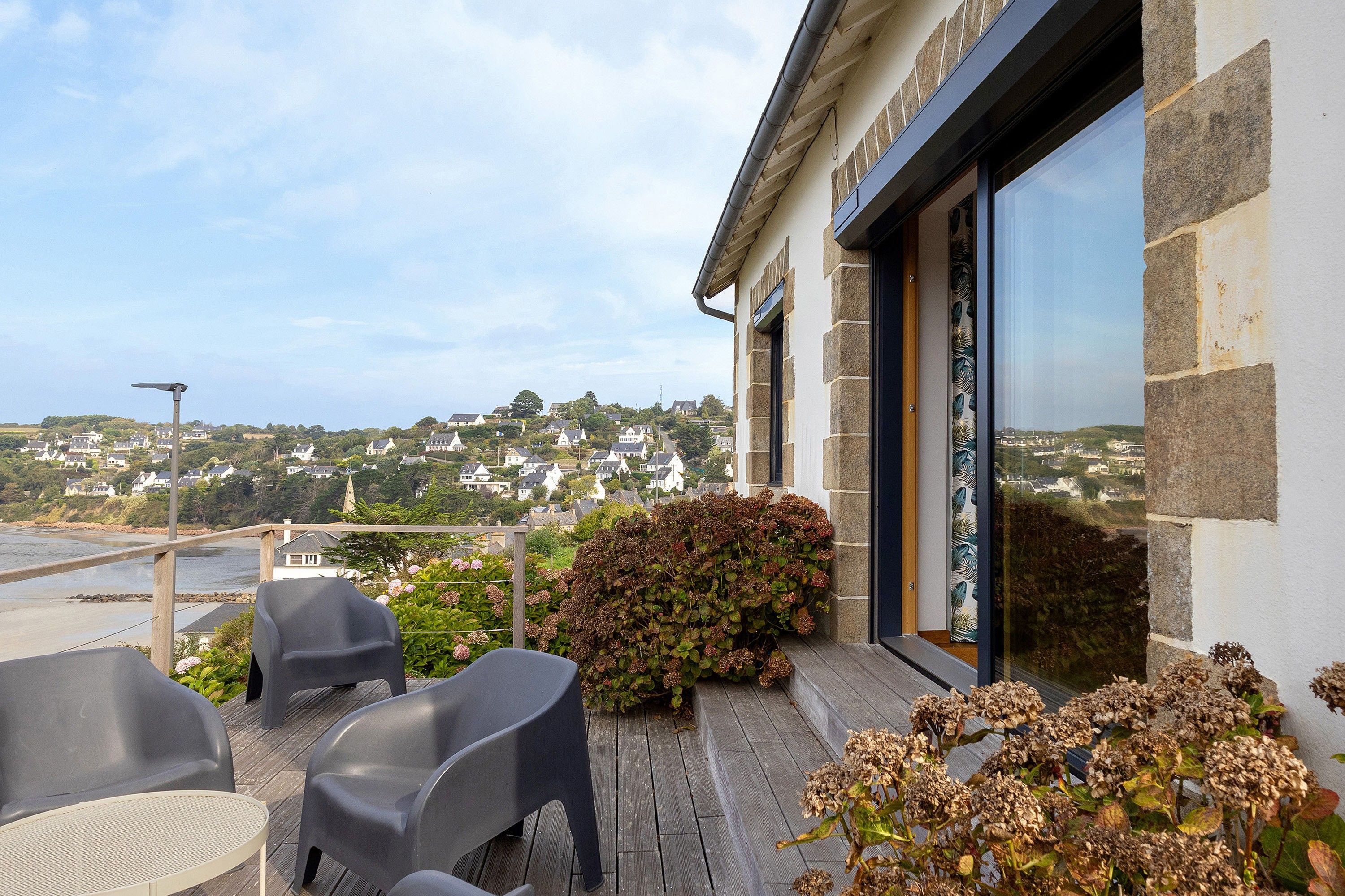 A balcony with outdoor seating, a view of a hillside town, and a building with stone and glass features.