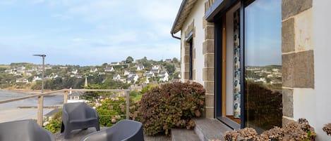 A balcony with outdoor seating, a view of a hillside town, and a building with stone and glass features.