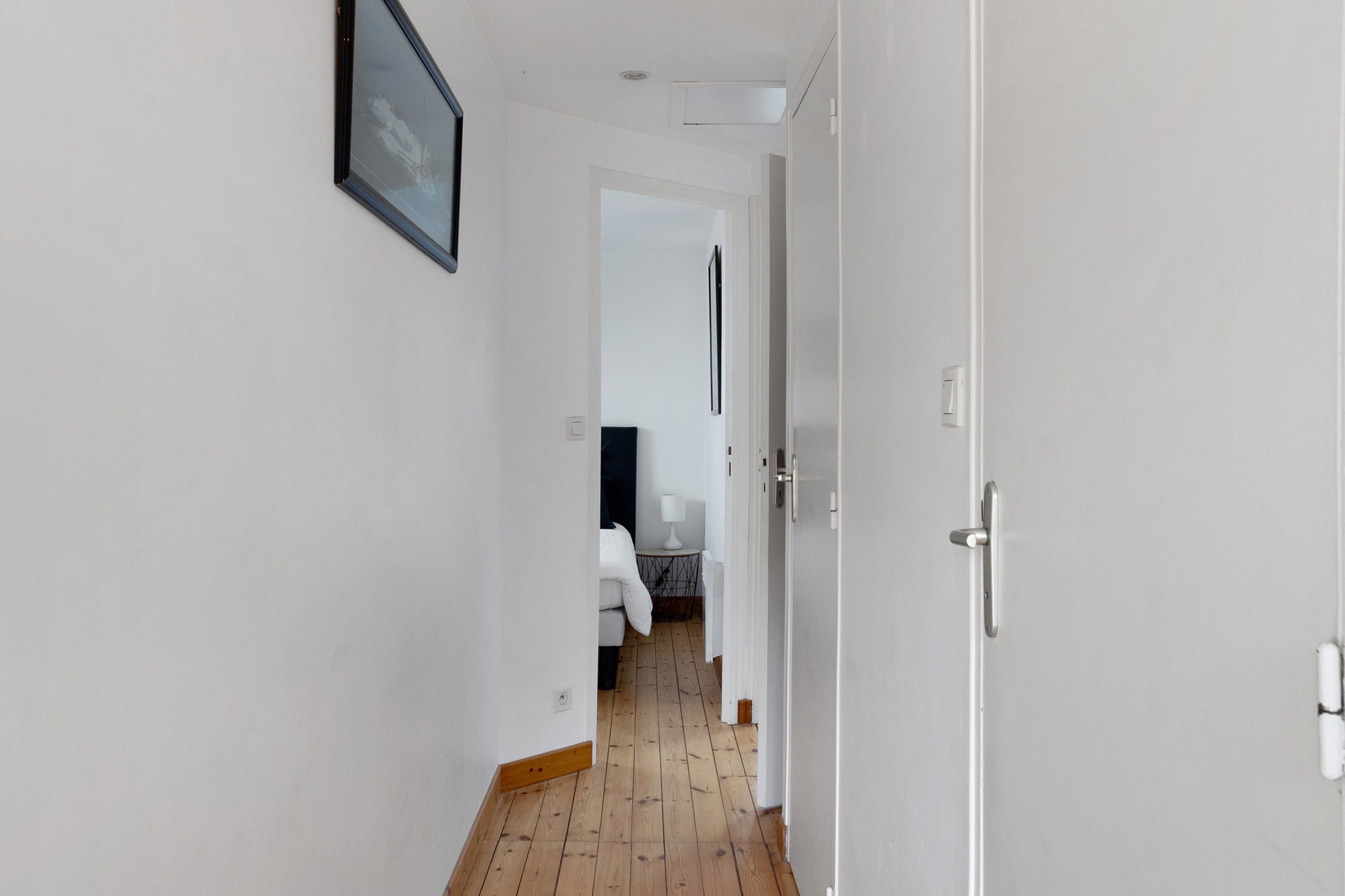 A hallway with a white door, a framed picture on the wall, and a glimpse of a room with a bed and a table.
