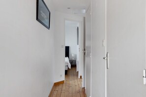 Un pasillo con una puerta blanca, un cuadro en la pared y una vista de una habitación con cama y mesa.
