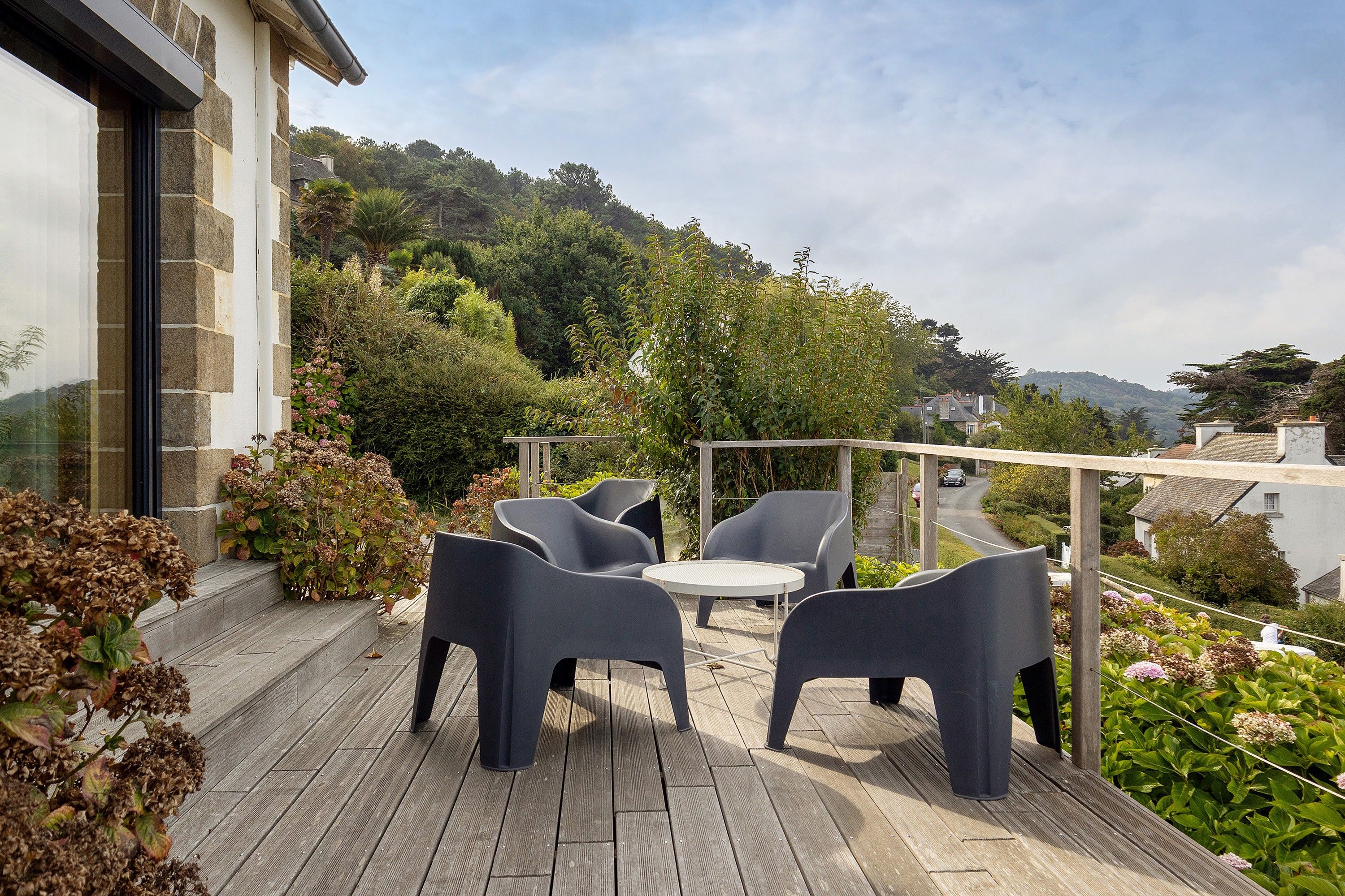 Un patio al aire libre con muebles modernos, rodeado de abundante follaje y vistas a una colina.