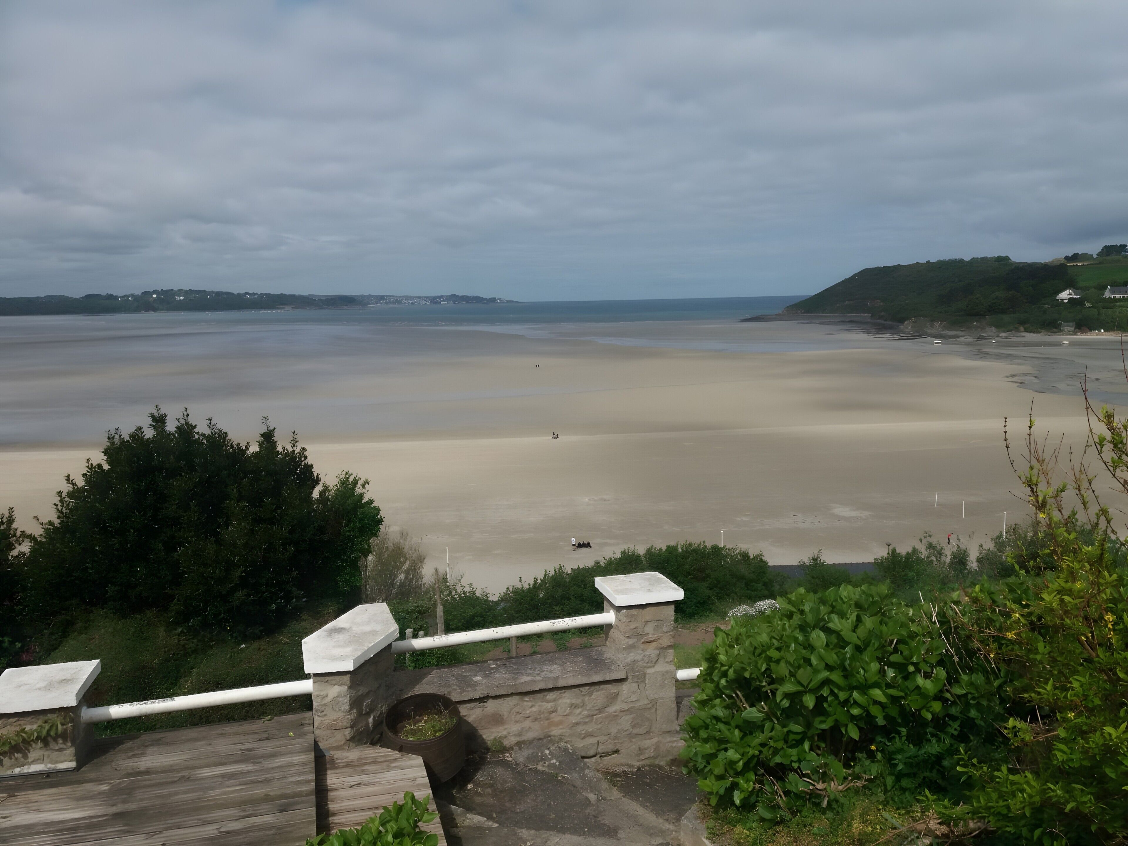 Blick auf die Küste mit einem Sandstrand, einer Steinmauer und viel Grün.