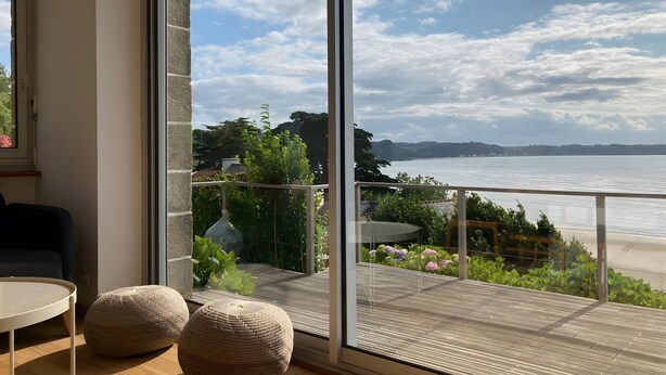 Una habitación con un gran ventanal que da a un balcón con vista al mar y al follaje.
