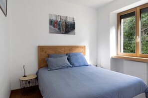 Un dormitorio con cabecera de madera, cama con ropa de cama azul, una mesita redonda con lámpara y ventana con vista a follaje.
