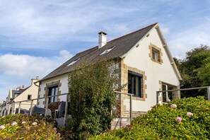 A two-story house with a sloped roof, stone facade, and a garden with various plants and flowers. - Holiday Home 'Maison Ty Bastian, 100 M²' with Sea View, Private Terrace and Wi-Fi (Saint-Michel-en-Grève)
