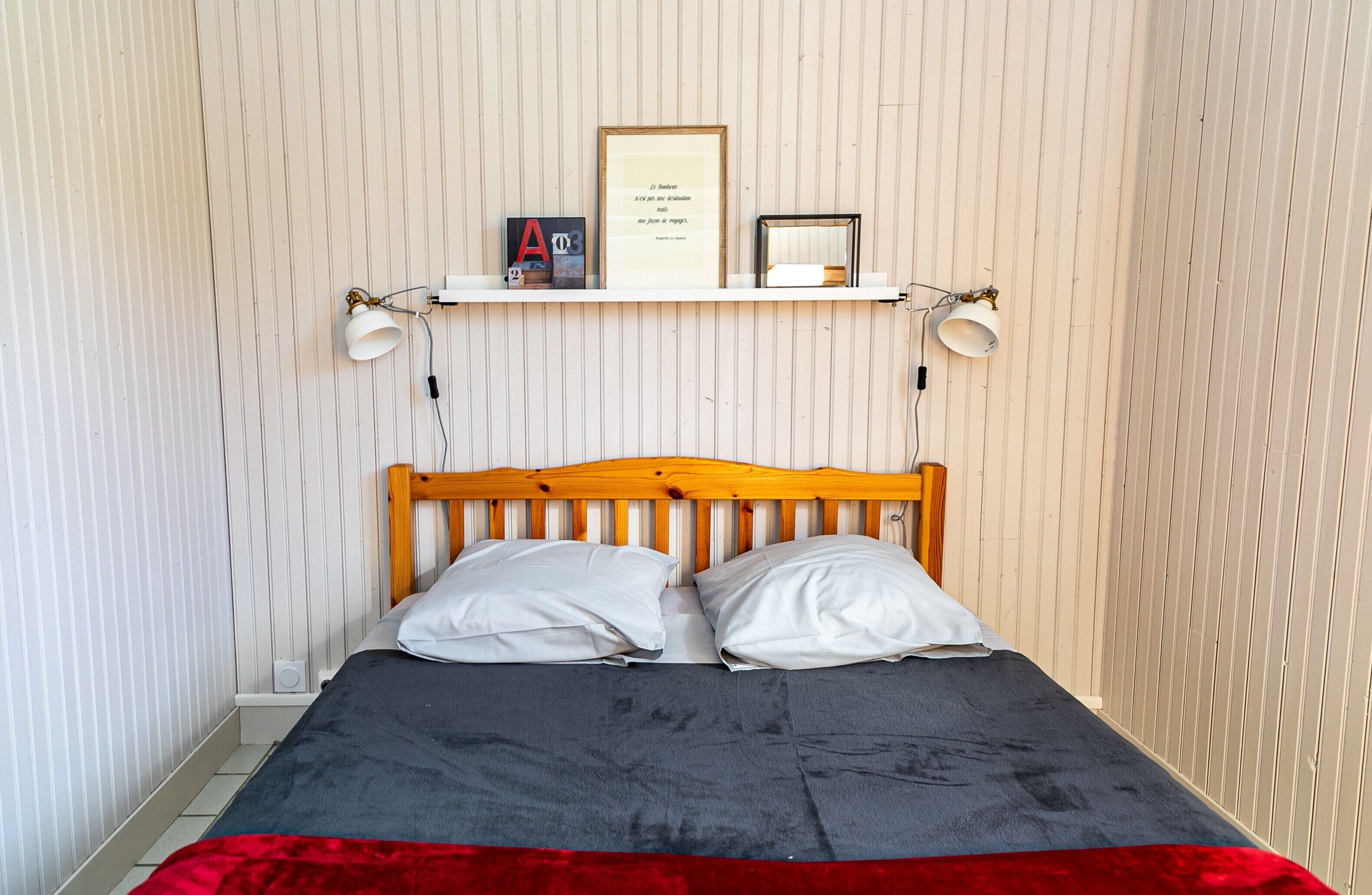 Een bed met een houten hoofdbord, twee witte kussens, een rode deken, een plank met boeken en een lijst met een citaat, plus twee wandlampen.