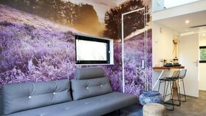Een moderne woonkamer met een grijze bank, een klein rond voetenbankje, een eethoek met een bar, en een groot muurdecoratie.