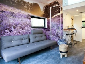 Ein modernes Wohnzimmer mit einem grauen Sofa, einem kleinen runden Hocker, einem Essbereich mit Bar und einem großen Wandbild.