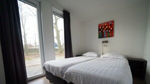 Ett hotellrum med en stor säng, ett sängbord, en liten avlastningsyta, ett fönster med gardiner och ett konstverk som hänger på väggen.
