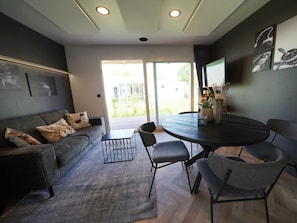 A modern living room with a grey sofa, a round table, and a large window. - Holiday Home in Kaatsheuvel With Terrace (Kaatsheuvel)
