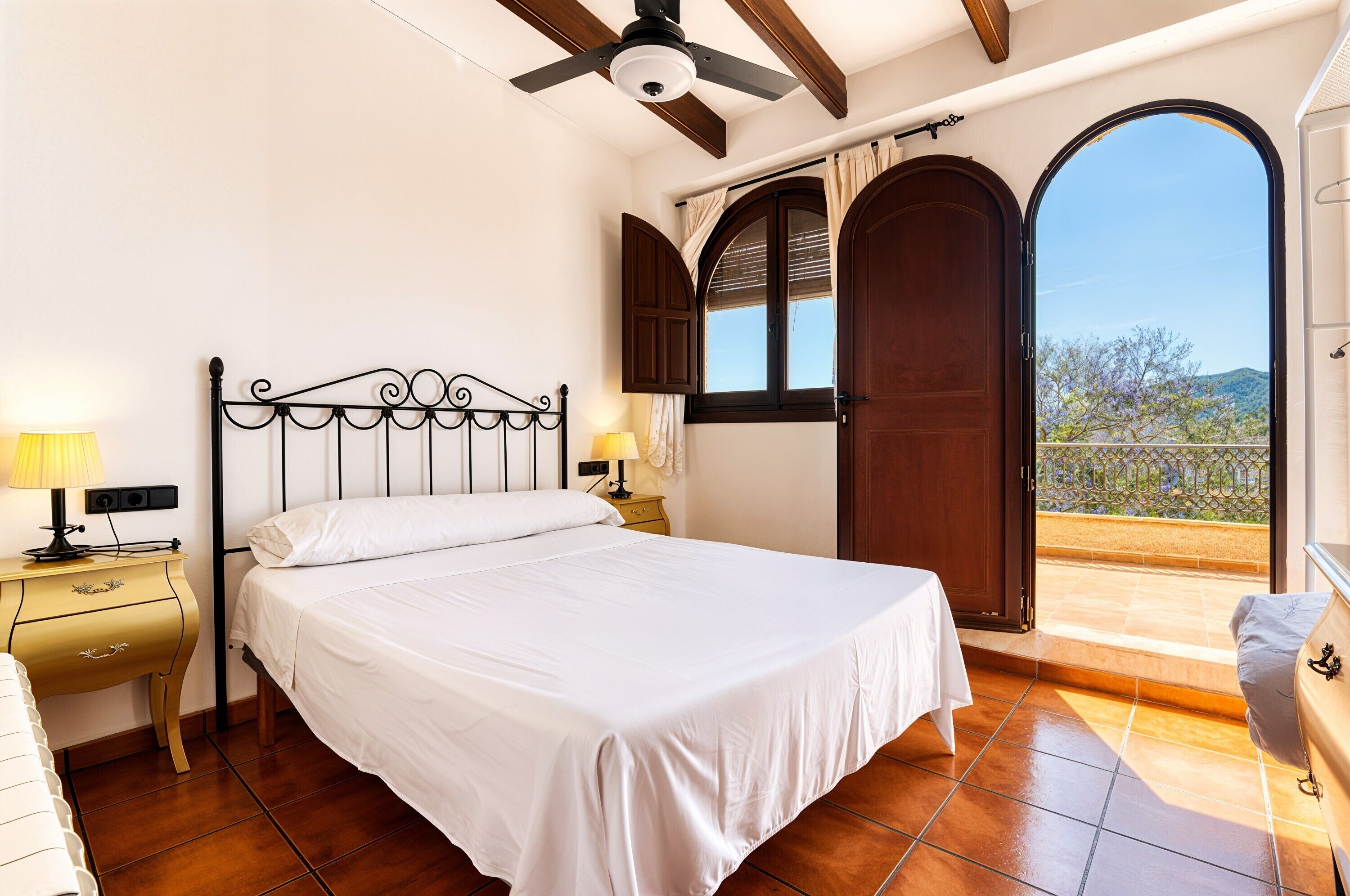 Une chambre à coucher avec un lit, deux tables de chevet avec des lampes, un ventilateur de plafond, une porte en bois et une vue sur l’extérieur.