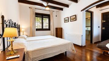 Une chambre d’hôtel comprenant un lit, une table de chevet avec une lampe, un ventilateur de plafond, une fenêtre avec des rideaux, un radiateur et une porte donnant sur une autre pièce.