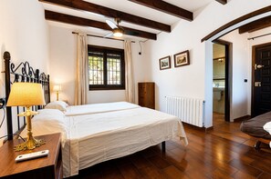 Habitación de hotel con cama, mesita de noche con lámpara, ventilador de techo, ventana con cortinas, radiador y puerta que da a otra habitación.