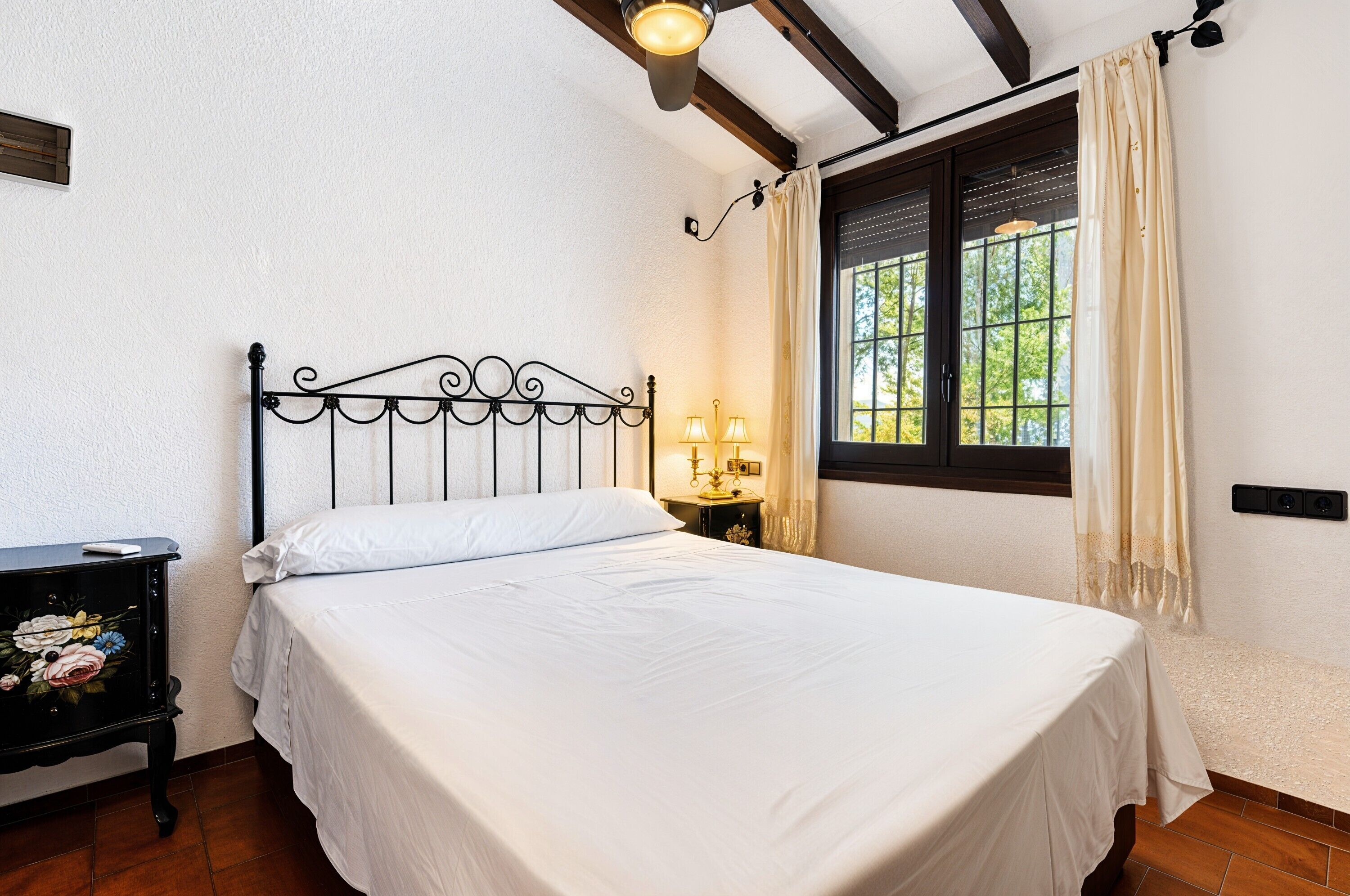 A neatly made bed with a headboard, a bedside table with a lamp, and a window with curtains.