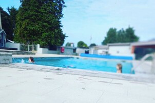 Zona de piscina con solárium empedrado, algunas personas nadando y árboles al fondo.