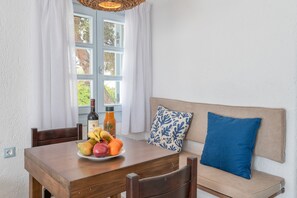 A dining table with a plate of fruit and two bottles, a wooden chair, a bench with a cushion, and a window with curtains. - Mykonian Dream Studio – Pool, Beach & Luxury (Mykonos)