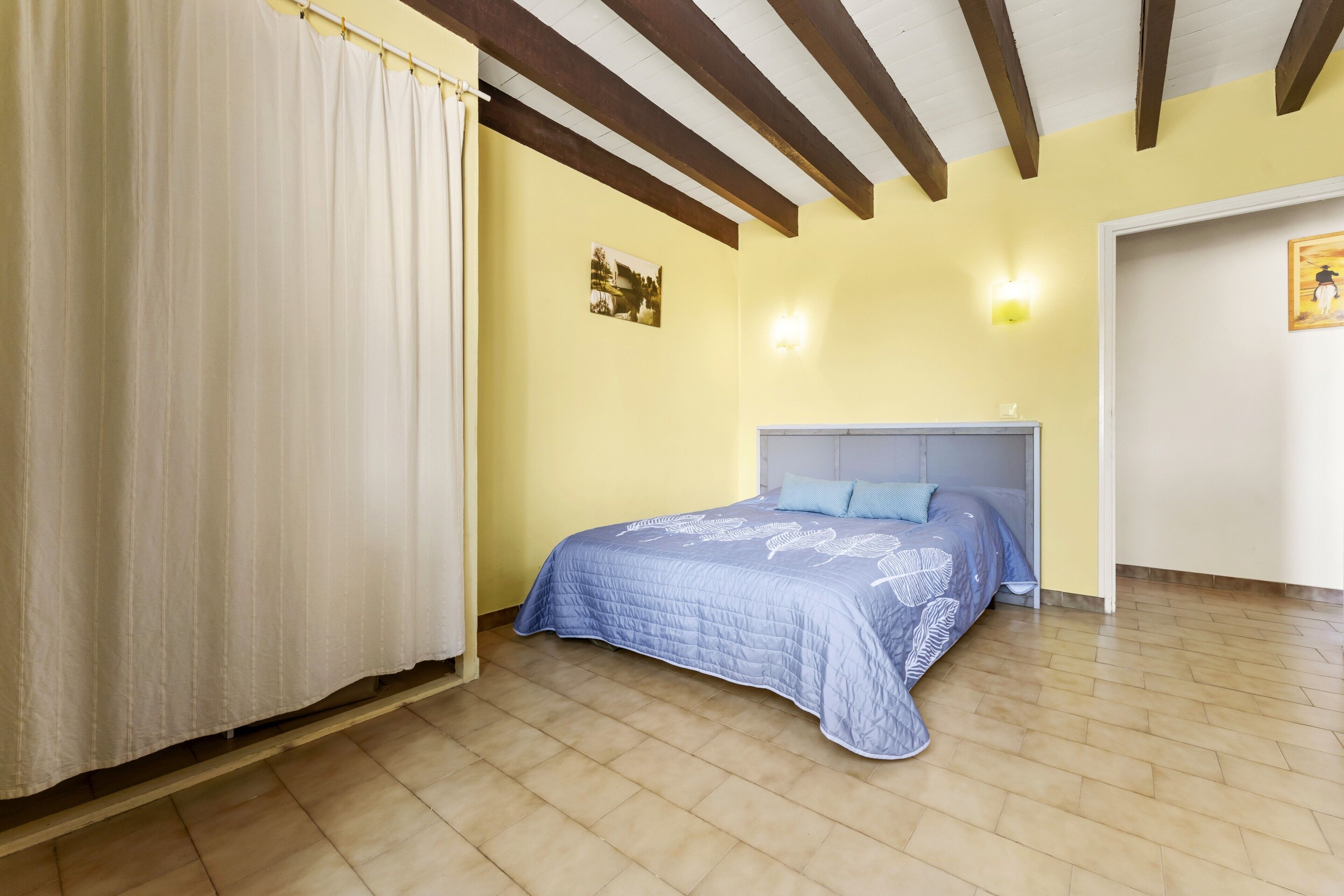 Un dormitorio con cama, cortina, lámpara de pared y un cuadro.