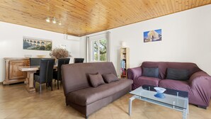 A living room with a brown sofa, a purple sofa, a glass coffee table, a wooden dining table with chairs, a bookshelf, and a wooden ceiling.