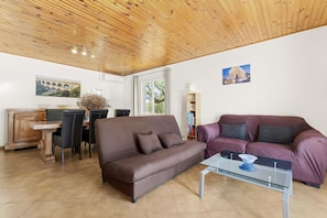 Una sala de estar con un sofá marrón, un sofá morado, una mesa de centro de vidrio, una mesa de comedor de madera con sillas, una estantería y un techo de madera.
