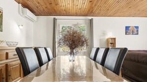 A dining area with a wooden table, black chairs, a vase with dried flowers, a bookshelf, and a brown sofa.