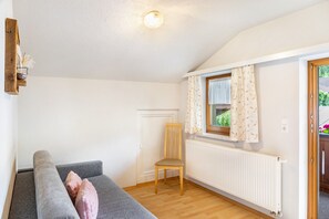 Ein Zimmer mit grauem Sofa, einem Holzstuhl, einem Fenster mit gemusterten Vorhängen, einer Heizung und einer Holztür.