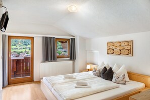 A bedroom with a large bed, a wooden headboard, a window with curtains, a view of greenery, and a wooden wall decoration.