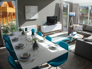 Zona pranzo con tavolo apparecchiato per quattro persone, divano, televisione e vista sull'esterno.