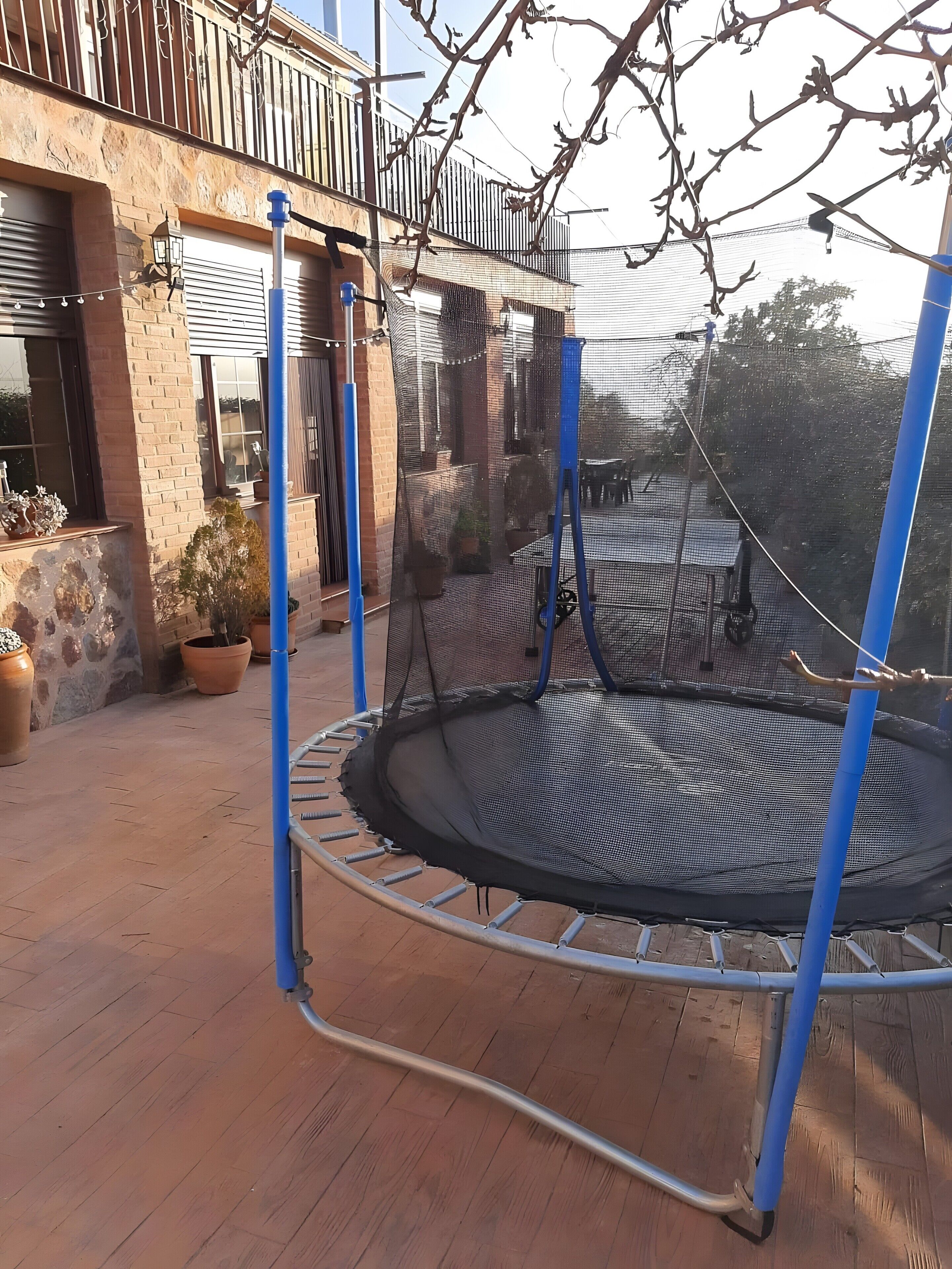 En trampolin med sikkerhedsnet, omgivet af en flisebelagt terrasse med plantekasser.
