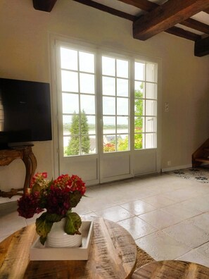 Una habitación con un gran ventanal, un televisor sobre un mueble bajo y un florero con flores sobre una mesa.