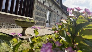 Un bâtiment traditionnel à colombages, doté d’une façade en pierre, d’un grand bac en pierre contenant des plantes et d’une zone herbeuse agrémentée de fleurs.