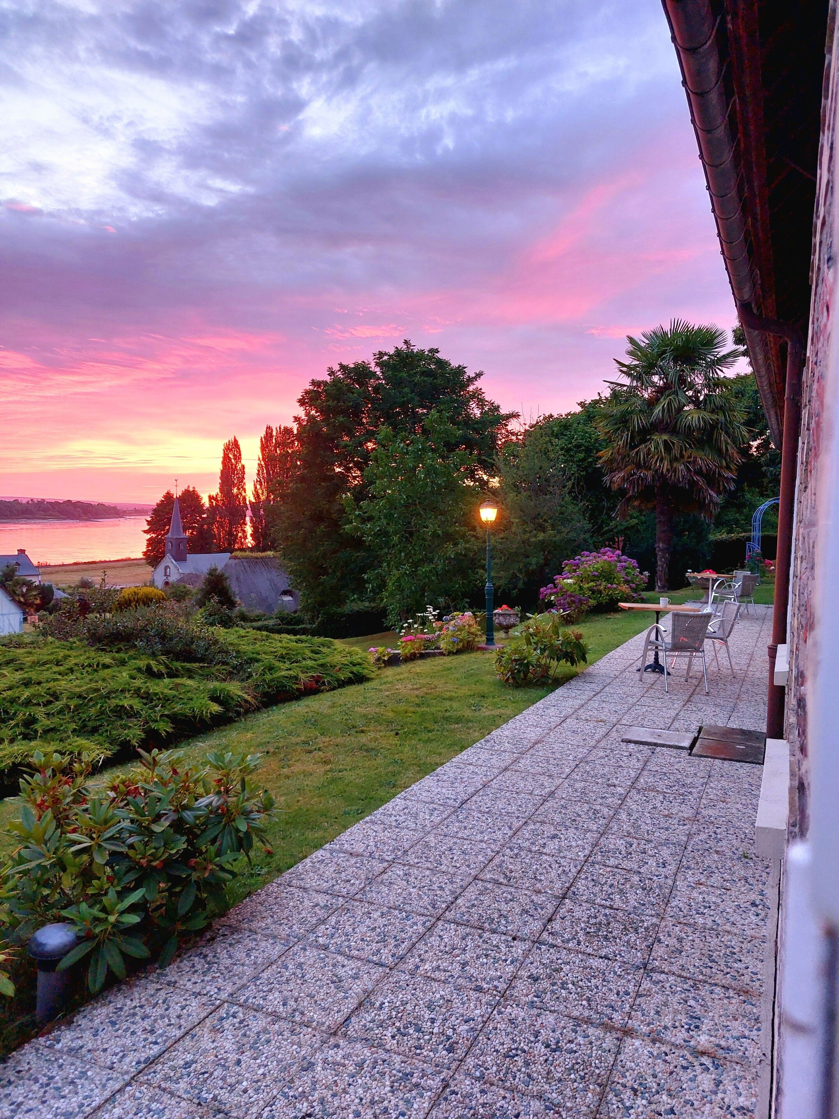 Une terrasse de jardin offrant une vue sur un coucher de soleil se reflétant sur une étendue d’eau, le tout entouré d’arbres et d’un lampadaire.