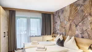 A hotel room with a large bed, two bedside tables, a headboard with a floral pattern, a window with sheer curtains, and a radiator. - Apartment Viktoria – Mountain Views, Terrace and Accessible Comfort (Ried im Zillertal)