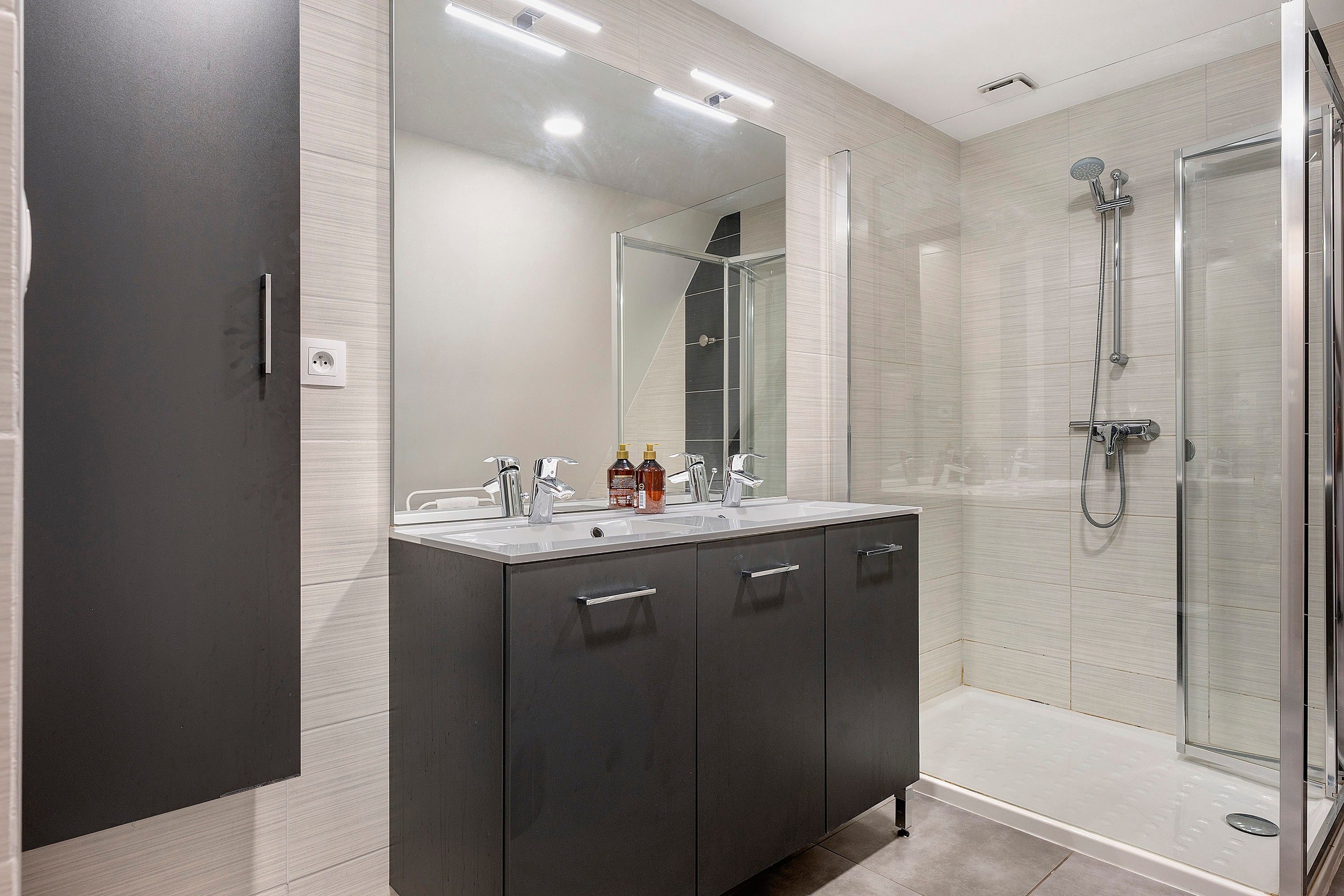 A modern bathroom with a dark vanity, a large mirror, a shower enclosure, and a white sink.