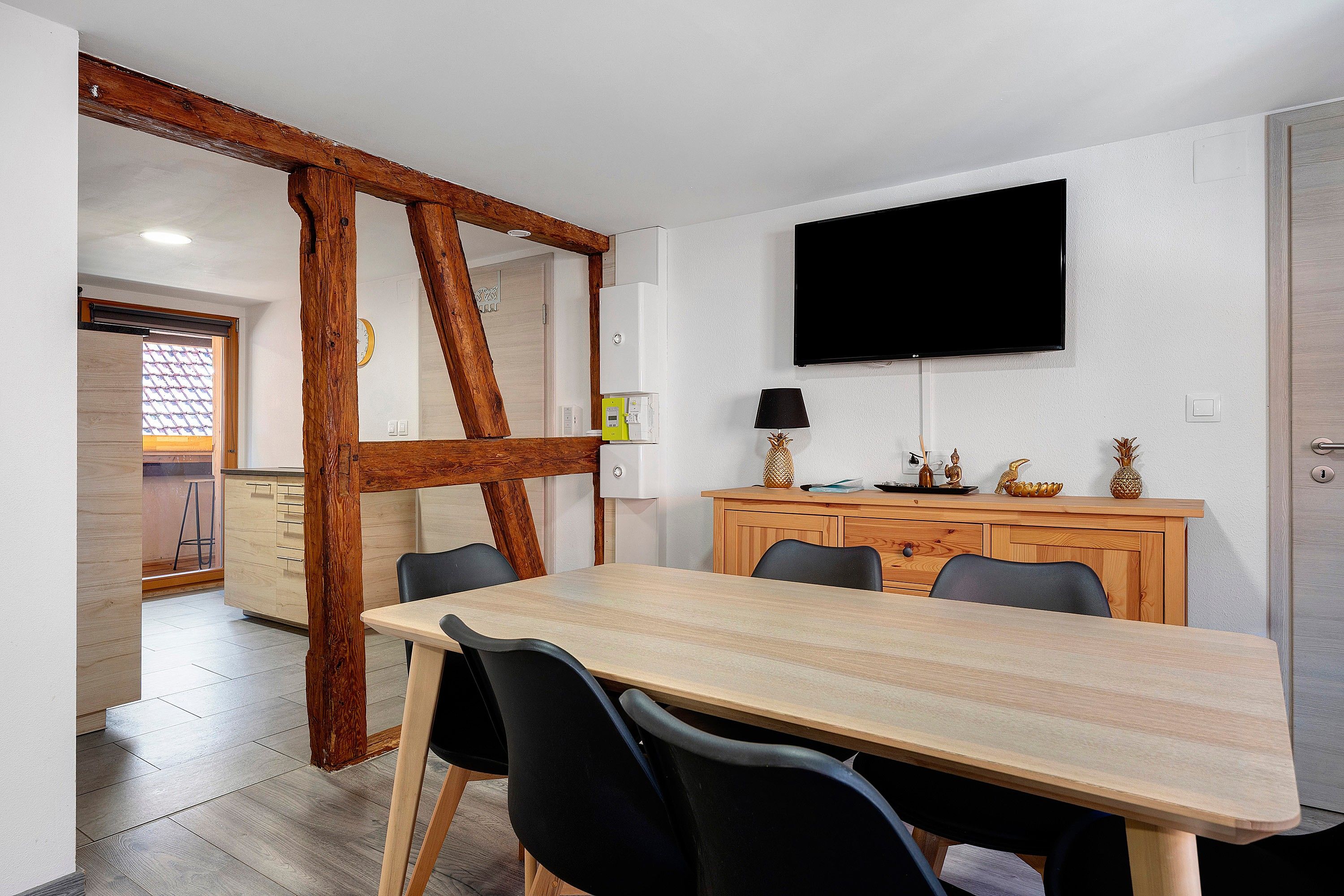 A dining area with a wooden table, chairs, a television mounted on the wall, and a wooden cabinet with decorative items.