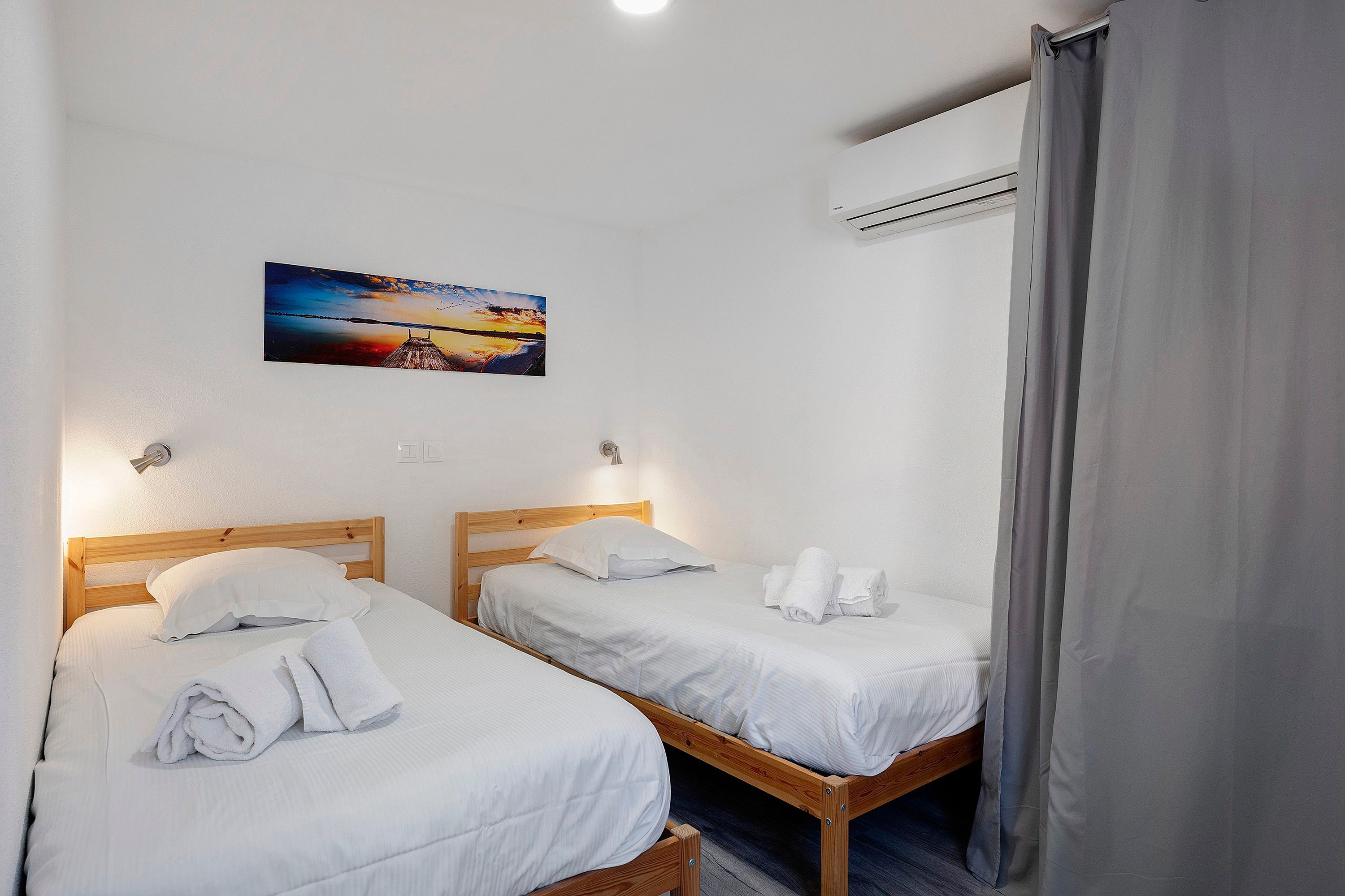 Ein Hotelzimmer mit zwei Einzelbetten, jeweils mit weißer Daunendecke und gerollten Handtüchern. An der Wand über den Betten hängt ein Gemälde eines Sonnenuntergangs.