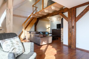 A loft-style room with exposed wooden beams, a sofa, a chair, and a staircase leading to an upper level.
