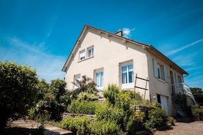 A two-story house with a stone staircase leading to the entrance, surrounded by lush greenery.