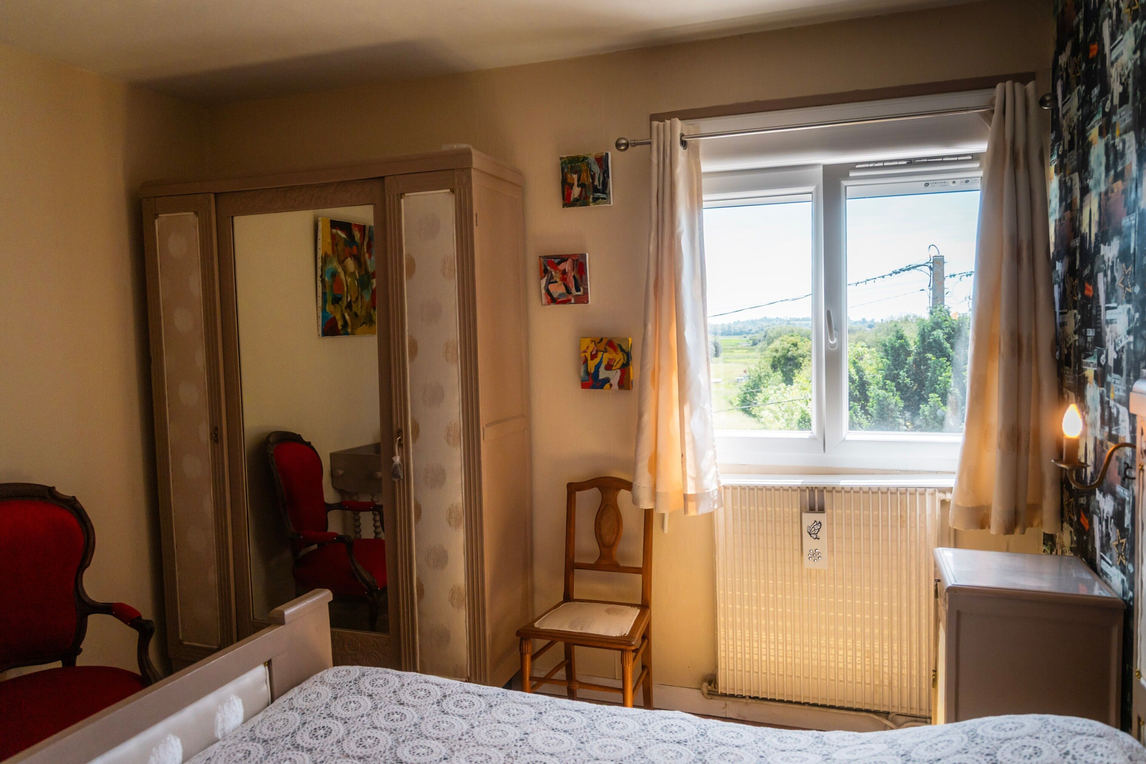 A bedroom with a bed, a chair, a wardrobe, a window with curtains, and artwork on the wall.
