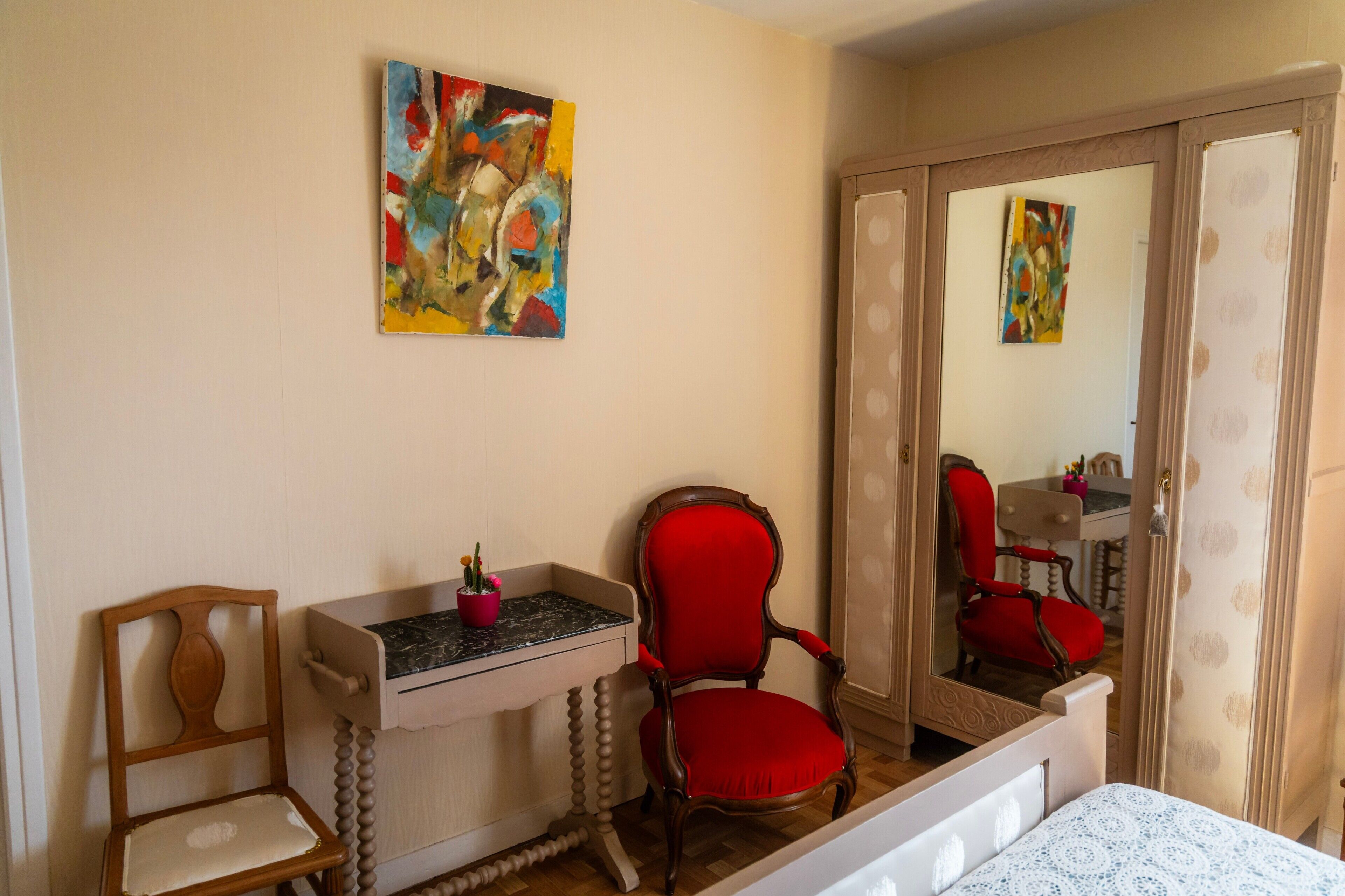 A room with a bed, a wooden chair, a desk with a vase, and a mirror reflecting a red chair and a dresser.