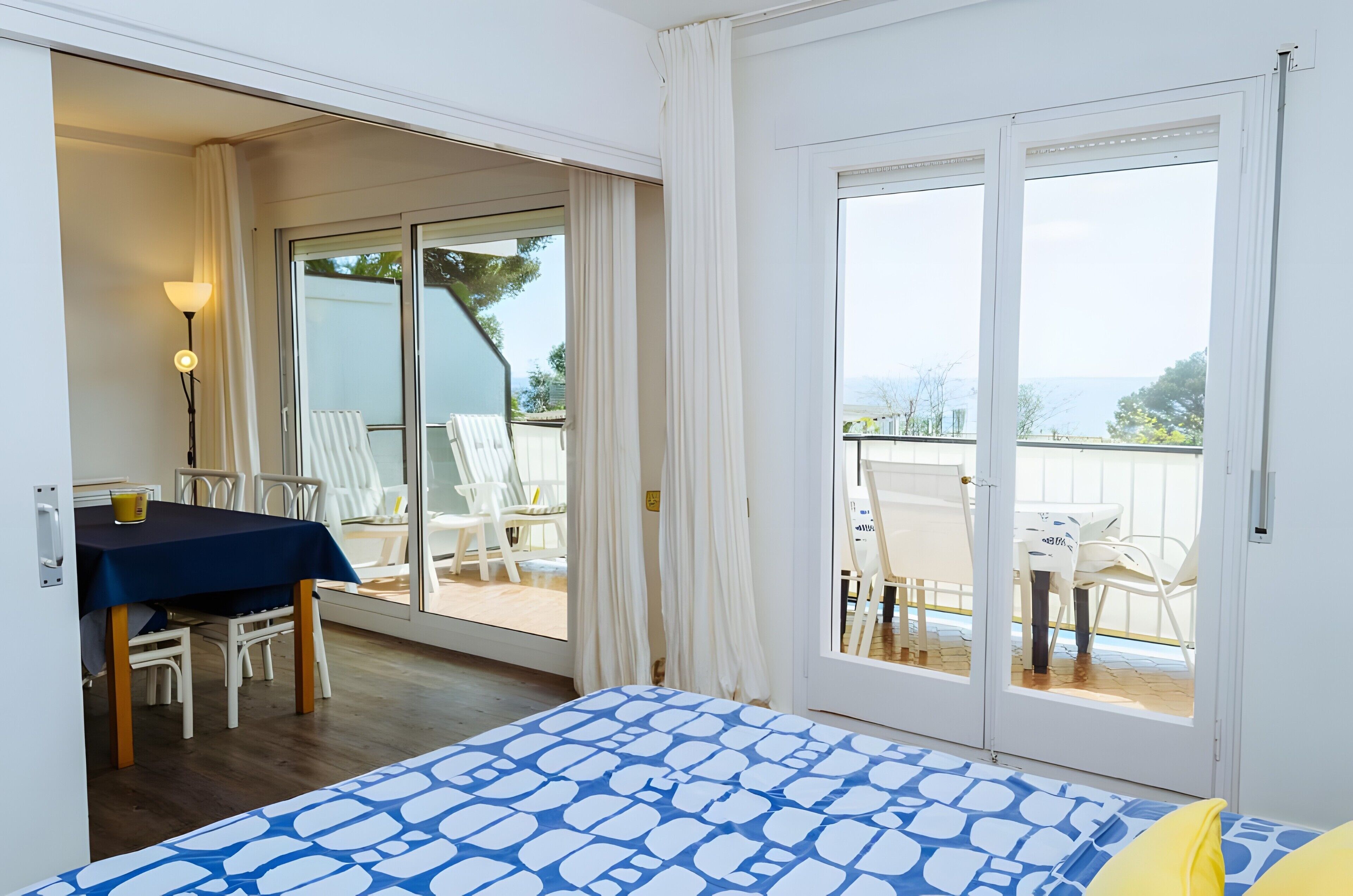 A bedroom with a bed, a table with chairs, a lamp, and a balcony with chairs and a table.
